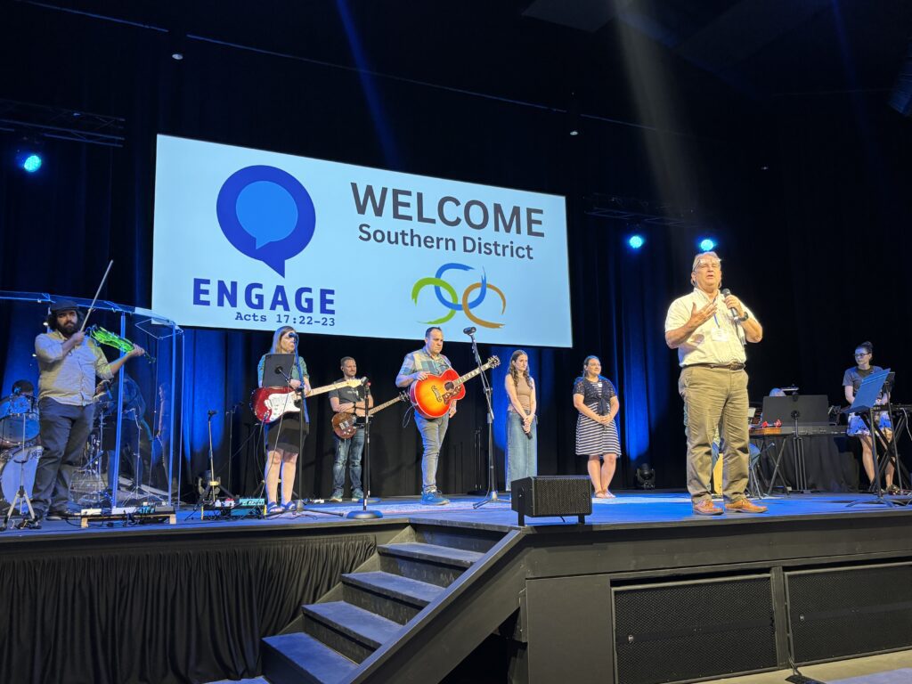 A man on stage extends a welcome while band members stand behind him.
