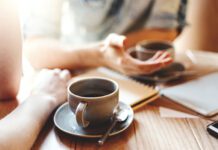 Patient and willing Friends talking at cafe table during coffee break. Unrecognizable colleagues discussing business issues, focus on coffee cup with saucer and teaspoon.