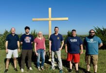 Confirming the call 5 men and 1 woman stand in front of a large wooden cross.