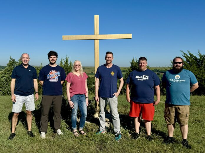 5 men and 1 woman stand in front of a large wooden cross.