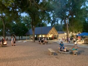 Early evening outdoors with people sitting around two fire pits and mingling around outside covered buildings.