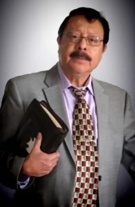 Man with dark hair, glasses, mustache, holding a Bible and wearing a gray suit and checkered tie.