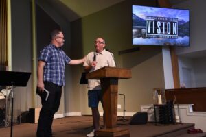 One pastor passes microphone to the other.