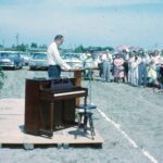 Man speaking outdoors beside piano.